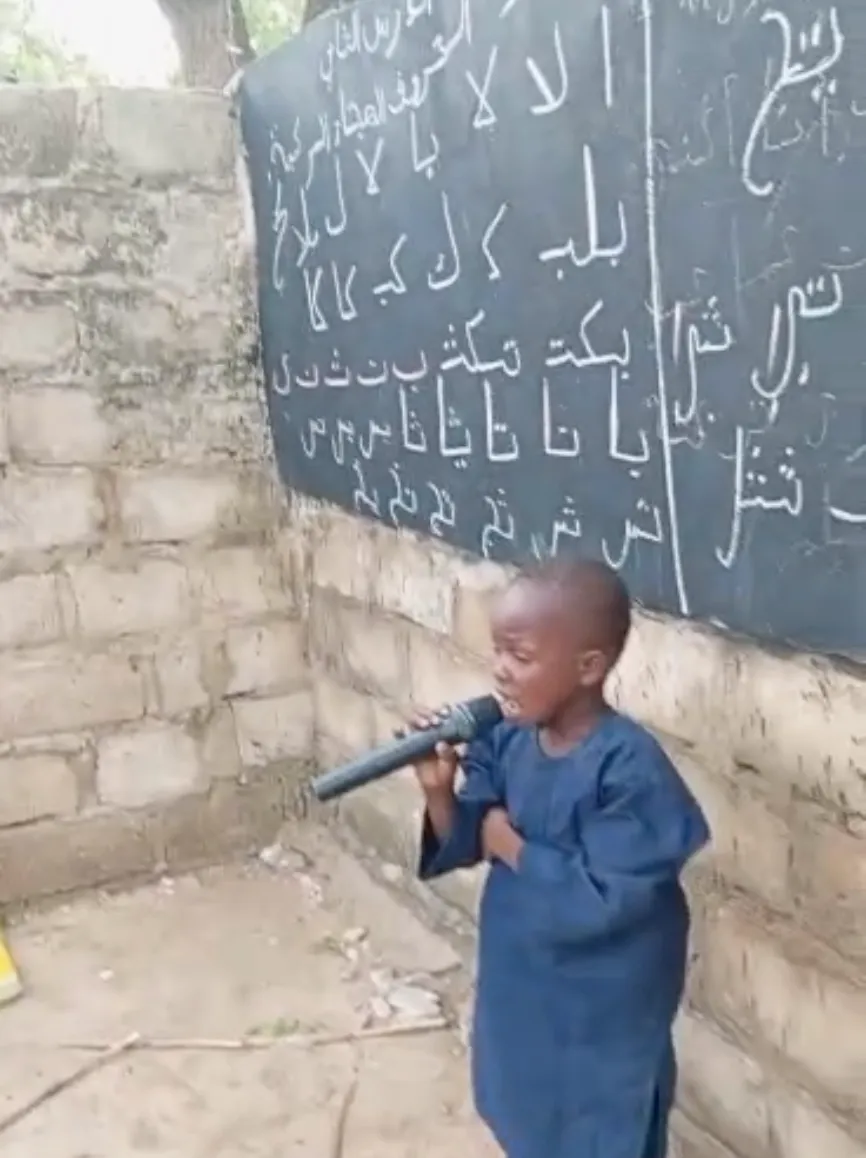 A young student reciting into a microphone beside the blackboard.